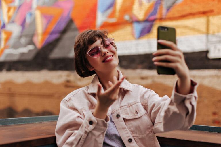 optimistic girl making selfie street woman with short brunette hair bright sunlasses denim beige jacket smiling outside