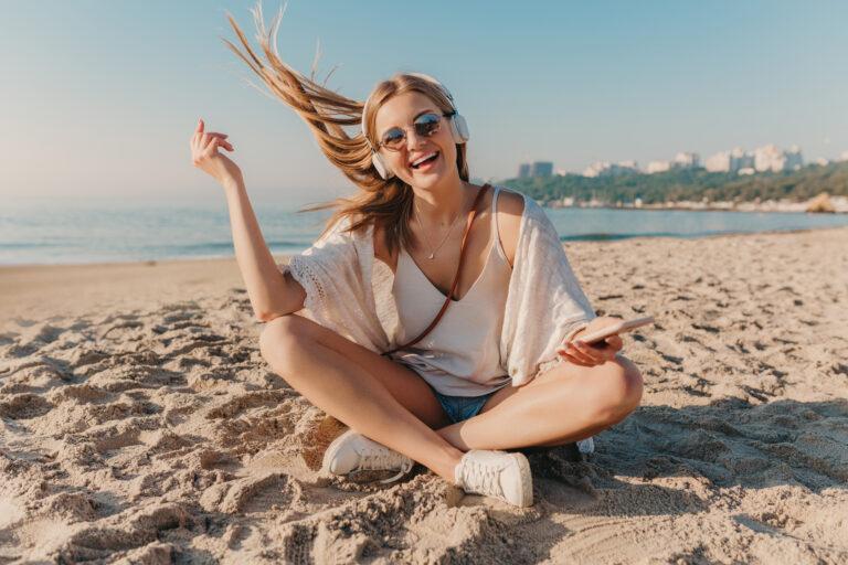 young attractive blond smiling woman sitting beach headphones listening music positive mood